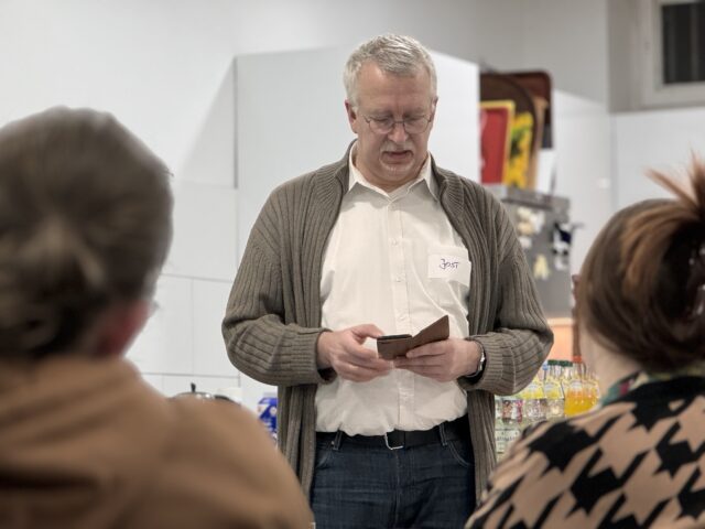 Ein Mann mit grauem Haar und Brille liest aus einem braunen Heft, während er in einem Raum steht, in dem im Vordergrund zwei Personen sitzen und hinter ihm auf einem Tresen Getränke stehen.
