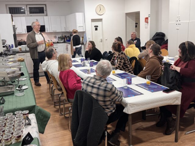 Eine Gruppe von Personen sitzt um zwei Tische in einer Küche oder einem Essbereich und hört einem Mann zu, der steht und spricht. Auf den Tischen stehen Speisen und Getränke, und die Atmosphäre wirkt locker und gesellig.