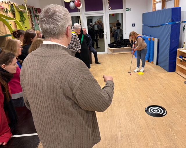 Eine Gruppe von Menschen sieht zu, wie eine Frau drinnen Minigolf spielt und auf eine schwarz-weiße Zielscheibe auf dem Boden zielt. Die Umgebung ist festlich mit Luftballons und Dekorationen an den Wänden.