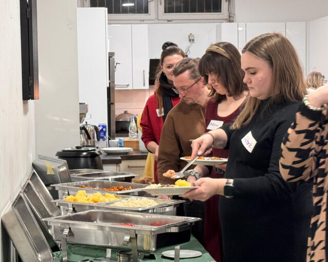 Eine Gruppe von Menschen steht in einer Reihe an einem Büffettisch und bedient sich in einer gut beleuchteten Küche an verschiedenen warmen Speisen. Die Personen scheinen sich darauf zu konzentrieren, ihre Teller mit Essen zu füllen.