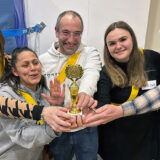 Fünf Personen stehen dicht beieinander, lächeln und halten eine kleine goldene Trophäe. Sie tragen gelbe Schärpen und wirken glücklich. Sie feiern als Gruppe in einem Raum in der Nähe eines Sportnetzes und eines Whiteboards.