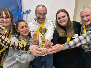 Fünf Personen stehen dicht beieinander, lächeln und halten eine kleine goldene Trophäe. Sie tragen gelbe Schärpen und wirken glücklich. Sie feiern als Gruppe in einem Raum in der Nähe eines Sportnetzes und eines Whiteboards.