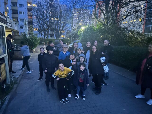 Eine Gruppe von Kindern und Erwachsenen versammelt sich in der Abenddämmerung in einem Wohngebiet mit Wohnhäusern und kahlen Bäumen im Hintergrund und lächelt. Einige Kinder halten einen Fußball in der Hand.