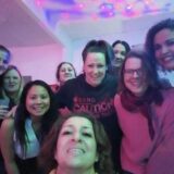 Eine Gruppe von zehn Frauen, die lächelnd und eng beieinander für ein Foto in einem Raum posieren, in dem bunte Lichter an der Decke eine festliche Atmosphäre schaffen.