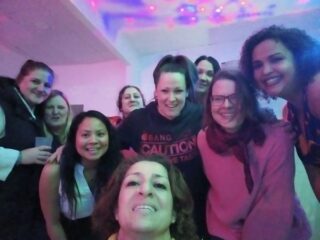 Eine Gruppe von zehn Frauen, die lächelnd und eng beieinander für ein Foto in einem Raum posieren, in dem bunte Lichter an der Decke eine festliche Atmosphäre schaffen.