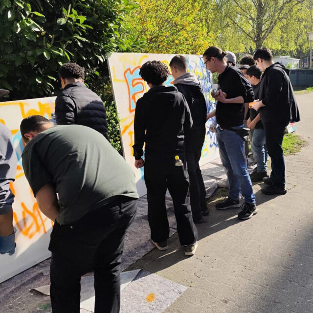 Eine Gruppe von Menschen steht im Freien und malt bunte Graffiti auf große Leinwände, die an Büsche gelehnt sind, während das Sonnenlicht durch die Bäume im Hintergrund fällt.
