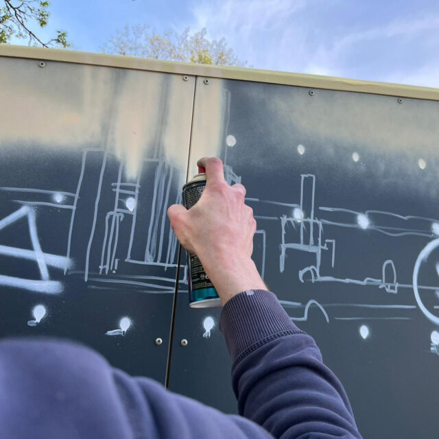 Eine Person sprüht eine weiße Stadtsilhouette mit Sternen oder Lichtern auf eine dunkle Wand oder Tafel im Freien, unter einem blauen Himmel, an dessen Spitze einige Bäume zu sehen sind.