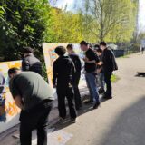 Eine Gruppe von Menschen steht im Freien neben weißen Tafeln mit bunten Graffiti, malt und zeichnet im hellen Sonnenlicht, umgeben von grünen Bäumen und Sträuchern.