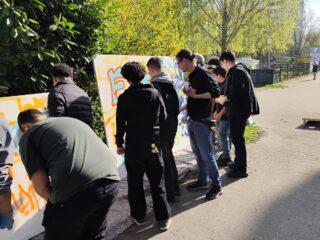 Eine Gruppe von Menschen steht im Freien neben weißen Tafeln mit bunten Graffiti, malt und zeichnet im hellen Sonnenlicht, umgeben von grünen Bäumen und Sträuchern.