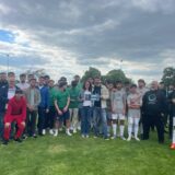 Eine große Gruppe von Menschen, darunter Fußballspieler in Uniformen und andere in Freizeitkleidung, posieren gemeinsam auf einer Wiese unter einem bewölkten Himmel. Einige halten eine Plakette oder eine Urkunde in der Mitte der Gruppe.