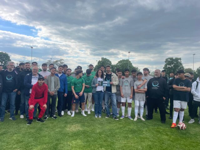 Eine große Gruppe von Menschen, darunter Fußballspieler in Uniformen und andere in Freizeitkleidung, posieren gemeinsam auf einer Wiese unter einem bewölkten Himmel. Einige halten eine Plakette oder eine Urkunde in der Mitte der Gruppe.