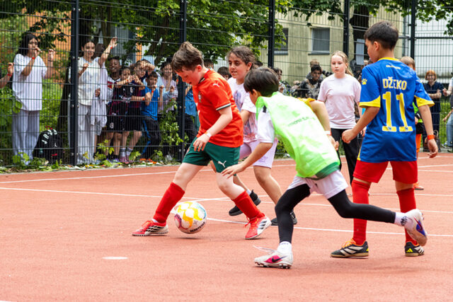 Kinder spielen ein Fußballspiel auf einem Platz im Freien. Mehrere Spieler in bunten Trikots jagen dem Ball nach, während eine Gruppe von Zuschauern hinter einem Metallzaun zuschaut und anfeuert. Im Hintergrund sind Bäume und Gebäude zu sehen.