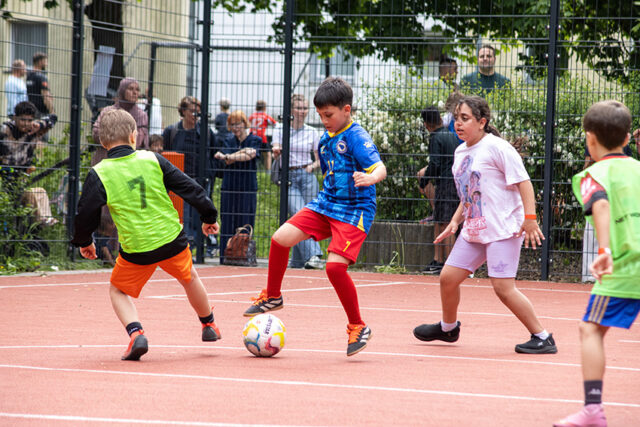 Vier Kinder spielen Fußball auf einem Platz im Freien. Ein Junge in einem blauen Trikot kontrolliert den Ball, während ein Junge in einer grünen Weste und zwei andere Kinder versuchen, ihn zu verteidigen. Die Zuschauer schauen hinter einem Zaun im Hintergrund zu.