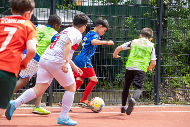 Eine Gruppe von Kindern in bunten Trikots spielt Fußball auf einem Platz im Freien. Ein Kind in Blau und Rot dribbelt den Ball, während andere in grünen, weißen und orangefarbenen Trikots hinterherlaufen und versuchen, ihn zu verteidigen.