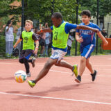 Eine Gruppe von Jungen spielt Fußball auf einem Platz im Freien. Ein Junge in einem grünen Lätzchen ist dabei, den Ball zu schießen, während andere neben ihm herlaufen. Die Zuschauer schauen hinter einem Zaun im Hintergrund zu.