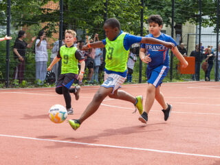 Eine Gruppe von Jungen spielt Fußball auf einem Platz im Freien. Ein Junge in einem grünen Lätzchen ist dabei, den Ball zu schießen, während andere neben ihm herlaufen. Die Zuschauer schauen hinter einem Zaun im Hintergrund zu.
