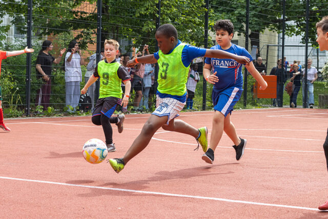 Eine Gruppe von Jungen spielt Fußball auf einem Platz im Freien. Ein Junge in einem grünen Lätzchen ist dabei, den Ball zu schießen, während andere neben ihm herlaufen. Die Zuschauer schauen hinter einem Zaun im Hintergrund zu.