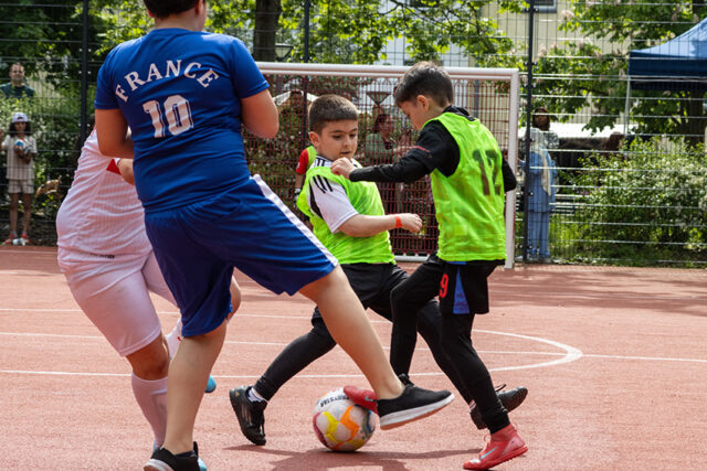 Drei Jungen spielen Fußball auf einem Platz im Freien. Zwei in grünen Westen kämpfen um den Ball, während ein Spieler in einem blauen France 10-Trikot und ein Spieler in Weiß versuchen, die Kontrolle zu übernehmen. Im Hintergrund sehen Zuschauer zu.