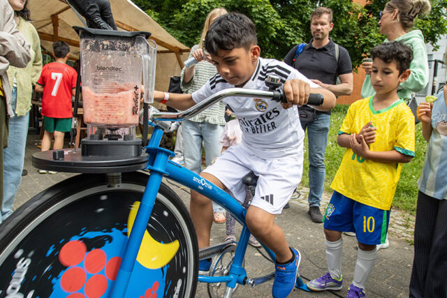 Ein Junge im Real-Madrid-Trikot tritt in die Pedale eines Standfahrrads, das einen Mixer antreibt. Er wird dabei von einem anderen Jungen im Brasilien-Trikot und anderen Leuten bei einer Veranstaltung im Freien beobachtet. Der Mixer ist mit rosa Smoothie-Zutaten gefüllt.