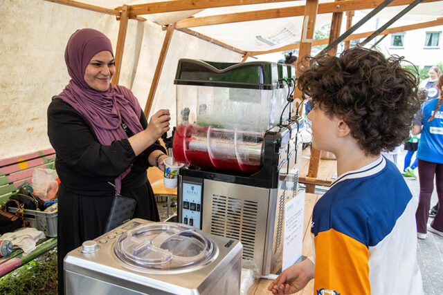Eine Frau in einem lila Hidschab serviert einem Jungen an einem Stand im Freien ein rotes Slush-Getränk. Der Junge, der ein weiß-blaues Hemd trägt, sieht zu, wie das Getränk aus einer Maschine unter einem Zelt ausgeschenkt wird.