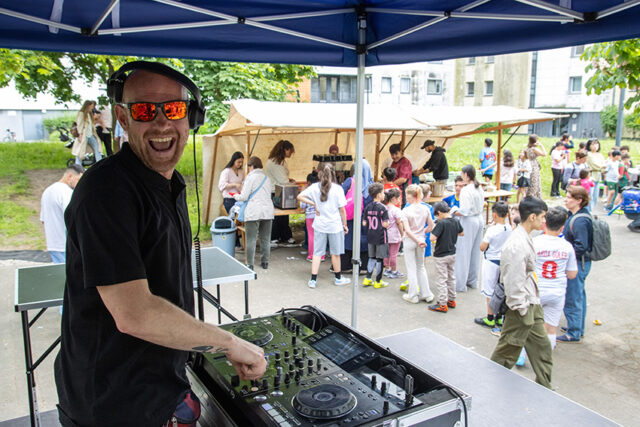 Ein DJ mit Kopfhörern und Sonnenbrille lächelt, während er bei einer Veranstaltung im Freien Musik auflegt, während sich im Hintergrund eine Gruppe von Kindern und Erwachsenen in der Nähe von Zelten versammelt.