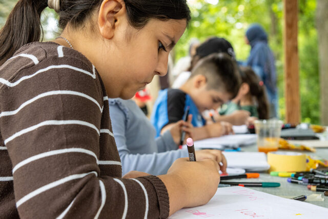 Ein junges Mädchen zeichnet mit einem Buntstift an einem Tisch im Freien, umgeben von anderen Kindern, die sich ebenfalls auf ihre Kunstwerke konzentrieren. Auf dem Tisch liegen Malutensilien und Papiere verstreut, und im Hintergrund sind Bäume zu sehen.