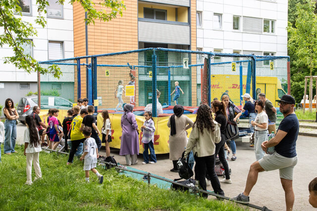 Eine Gruppe von Menschen, darunter Kinder und Erwachsene, versammelt sich draußen in der Nähe eines eingezäunten Trampolinbereichs, in dem Kinder spielen. Die Szene ist lebhaft, die Menschen stehen, unterhalten sich und schauen in der Nähe von Wohnhäusern zu.