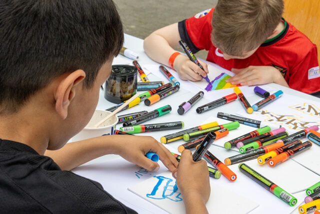 Zwei Kinder sitzen an einem Tisch und zeichnen mit bunten Markern auf weißem Papier. Verschiedene Stifte sind über den Tisch verstreut, und beide Kinder sind auf ihr Kunstwerk konzentriert.
