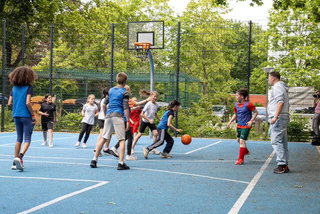 Eine Gruppe von Kindern spielt Basketball auf einem Platz im Freien, während ein Erwachsener an der Seitenlinie steht und zusieht. Die Szene ist lebhaft, mit Bäumen und einem eingezäunten Gelände im Hintergrund.