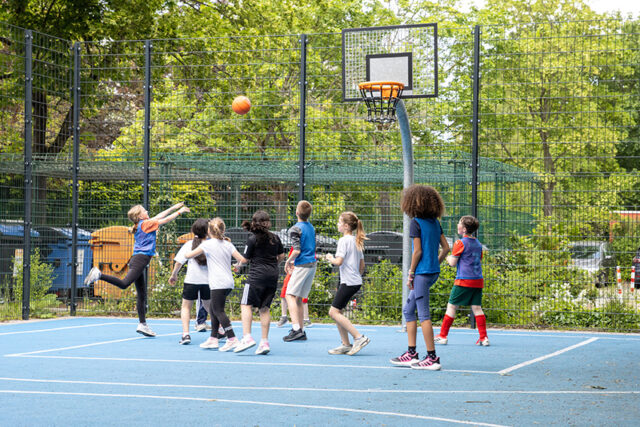 Eine Gruppe von Kindern spielt Basketball auf einem Platz im Freien. Ein Kind springt, um den Ball in Richtung Korb zu schießen, während andere zusehen oder sich auf den Abprall vorbereiten. Bäume und ein Zaun umgeben den Platz an einem sonnigen Tag.