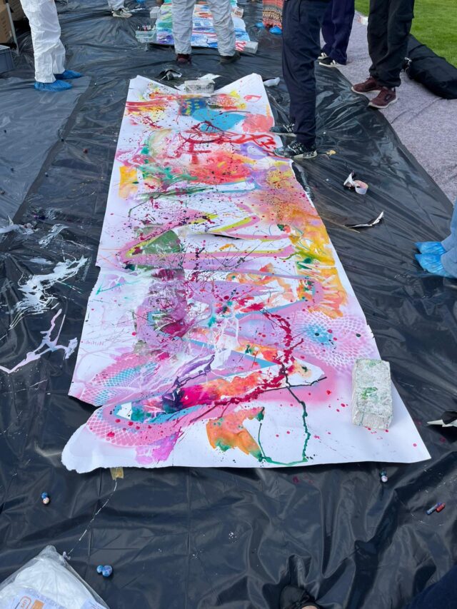 Ein großes abstraktes Gemälde mit leuchtenden Farbtupfern in Rosa, Orange, Grün und Violett ist draußen auf einer Plane ausgebreitet. Mehrere Personen in Schutzkleidung und mit Schuhüberzügen stehen um das Kunstwerk herum.