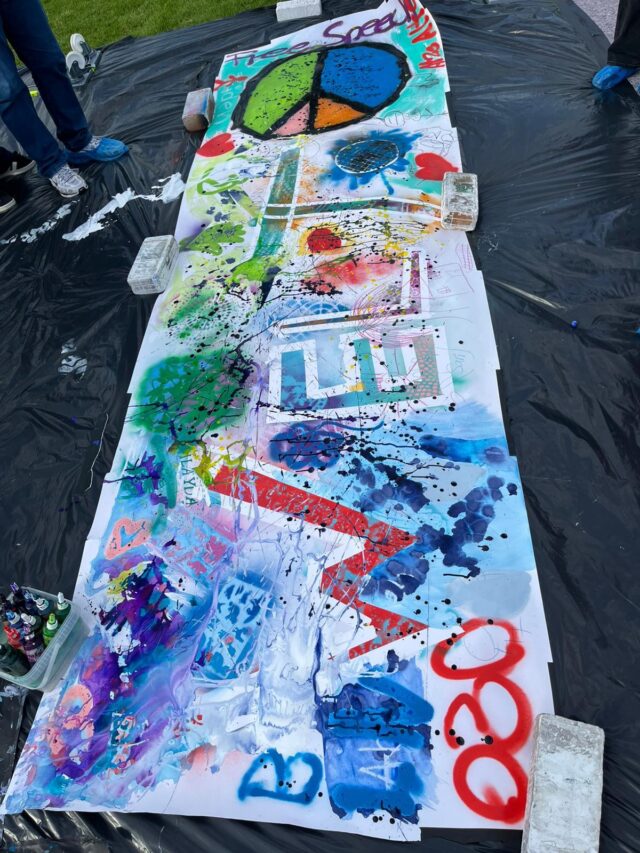 Ein langes Tuch, das mit bunten Motiven im Graffiti-Stil bedeckt ist – darunter ein großes Friedenszeichen, Herzen, Symbole und verschiedene Sprühfarbenmuster –, liegt draußen auf einer schwarzen Plane und wird an den Ecken und Rändern mit Ziegelsteinen beschwert.