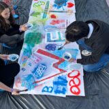 Mehrere Menschen knien um eine große weiße Leinwand herum, die auf dem Boden liegt, und malen bunte Muster und Symbole darauf, darunter ein Friedenszeichen, Herzen und die Zahlen „2020“. Die Leinwand liegt auf einer schwarzen Plastikplane.