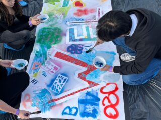 Mehrere Menschen knien um eine große weiße Leinwand herum, die auf dem Boden liegt, und malen bunte Muster und Symbole darauf, darunter ein Friedenszeichen, Herzen und die Zahlen „2020“. Die Leinwand liegt auf einer schwarzen Plastikplane.