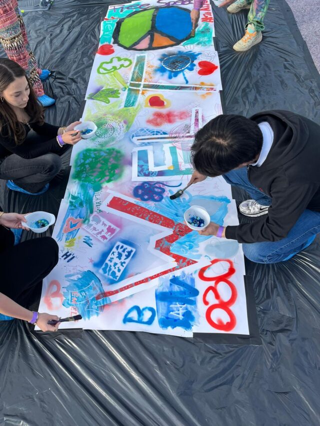 Mehrere Menschen knien um eine große weiße Leinwand herum, die auf dem Boden liegt, und malen bunte Muster und Symbole darauf, darunter ein Friedenszeichen, Herzen und die Zahlen „2020“. Die Leinwand liegt auf einer schwarzen Plastikplane.