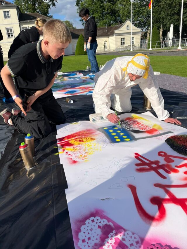 Zwei Personen knien draußen auf einer Plane und gestalten mit Sprühfarbe farbenfrohe Kunstwerke auf großen Papierbögen; eine ist schwarz gekleidet, die andere trägt einen weißen Schutzanzug. Sprühdosen sind zu sehen, und im Hintergrund schauen andere zu.