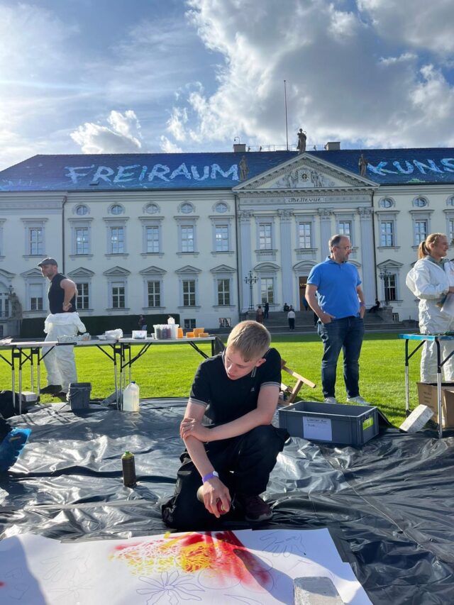 Vor einem großen historischen Gebäude kniet ein Junge auf einer Plane und arbeitet mit Sprühfarbe an einem Kunstwerk. In der Nähe befinden sich weitere Personen und Malutensilien; auf dem Dach des Gebäudes ist „FREIRAUM“ zu sehen. Der Himmel ist teilweise bewölkt.
