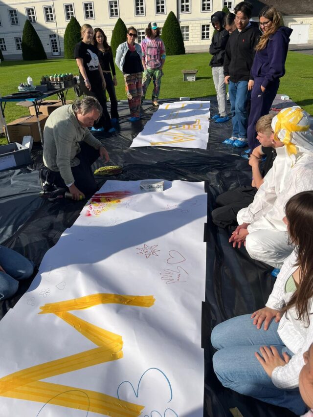 Eine Gruppe von Menschen, hauptsächlich Jugendliche, sitzt und steht draußen auf dem Rasen um große Papierbögen herum und zeichnet und malt im Sonnenlicht mit leuchtenden Farben. Ein Kursleiter kniet neben den Kunstwerken und leitet die Aktivität an.