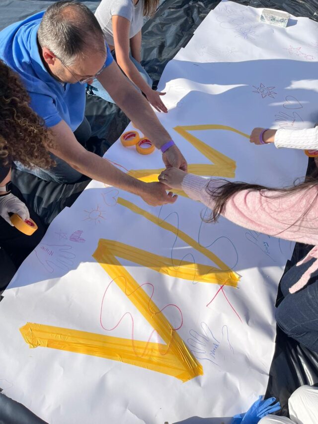 Vier Personen arbeiten gemeinsam an einem großen weißen Blatt Papier, kleben gelbe Linien auf und zeichnen bunte Formen, Handumrisse und Kritzeleien. Die Aktivität wirkt gemeinschaftlich und kreativ und findet im Freien bei strahlendem Sonnenschein statt.