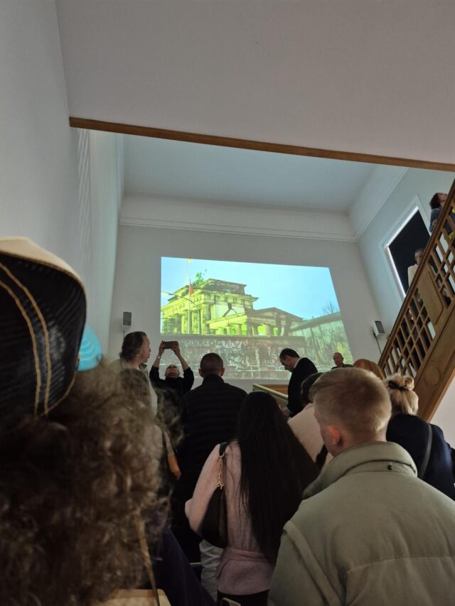 Eine Gruppe von Menschen steht in einem Treppenhaus und blickt auf die Projektion eines großen Gebäudes mit einer Flagge. Einige der Anwesenden machen Fotos oder Videos mit ihren Handys. Die Atmosphäre wirkt konzentriert und aufmerksam.