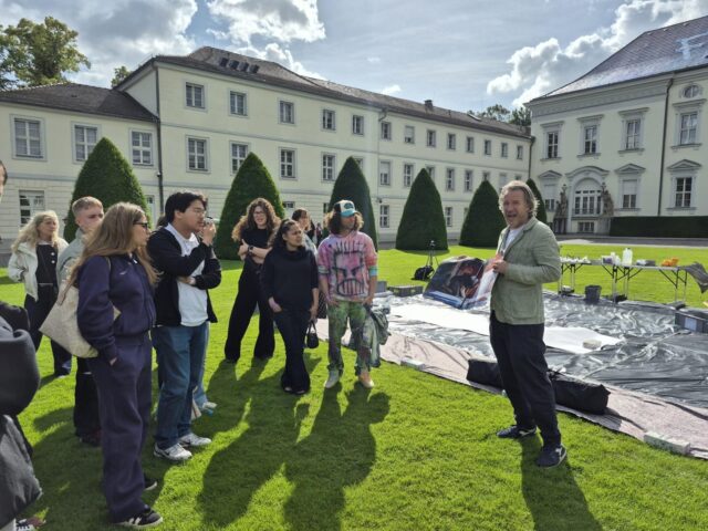Eine Gruppe von Menschen steht auf einer sonnigen Wiese vor einem großen historischen Gebäude und hört einem Mann zu, der spricht. Auf dem Rasen liegen Malutensilien verstreut, was auf einen Workshop oder einen Kurs im Freien hindeutet.