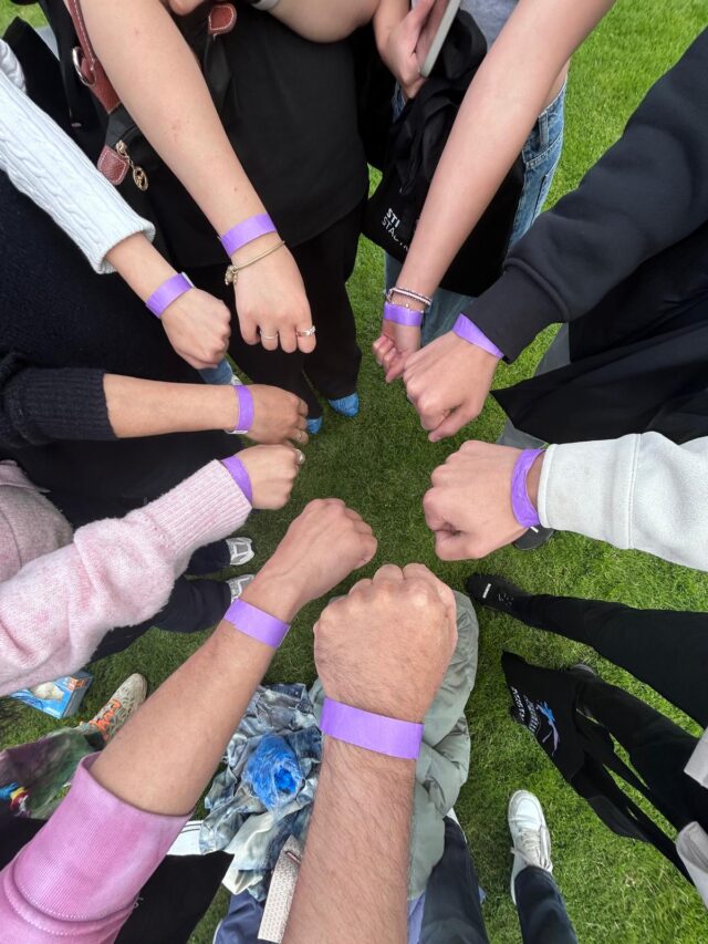 Eine Gruppe von Menschen, die im Freien im Kreis steht, reckt ihre Fäuste in die Höhe; jeder trägt ein lila Armband, im Hintergrund ist grünes Gras zu sehen.