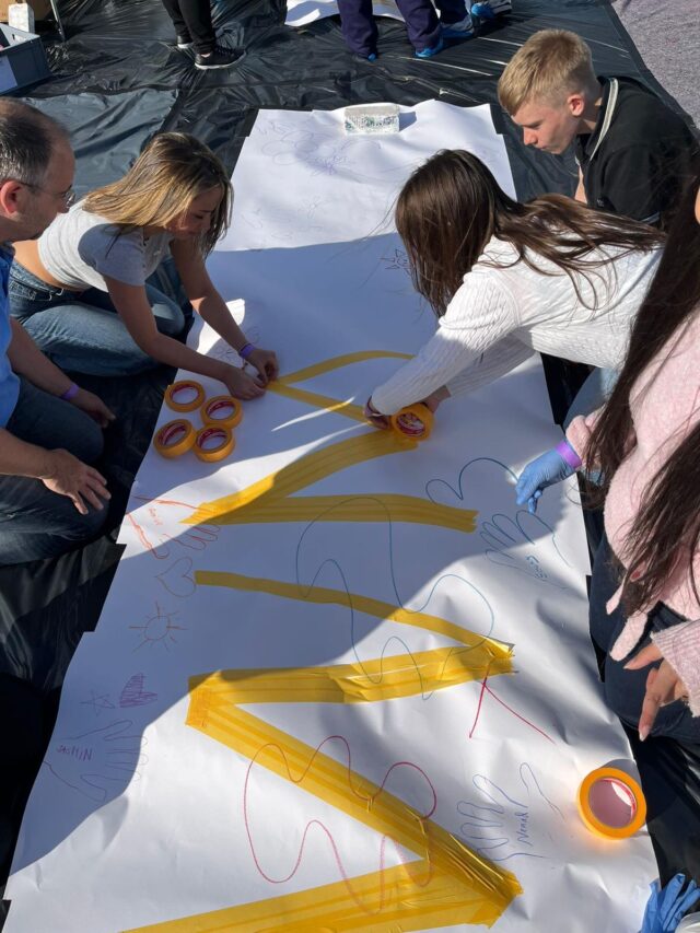 Mehrere Kinder und Erwachsene arbeiten gemeinsam an einem großen weißen Blatt Papier, befestigen es mit gelbem Klebeband und malen bunte Formen wie Hände, Herzen und Sonnen darauf. Die Aktivität findet an einem sonnigen Tag im Freien statt.
