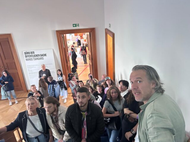 Eine Gruppe von Menschen steht und sitzt auf einer Treppe in einem Innenraum und blickt zur Kamera hinauf. Einige halten Handys oder Kameras in der Hand. Hinter ihnen sind ein Flur mit Menschen und ein Schild in deutscher Sprache an der Wand zu sehen.