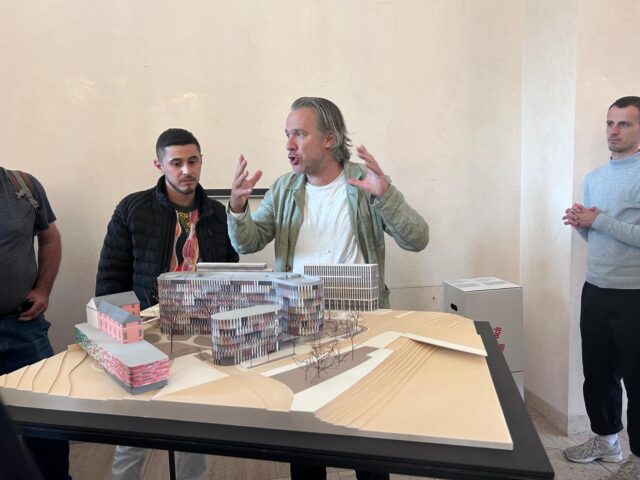 Ein Mann gestikuliert lebhaft, während er einer Gruppe von Menschen, die sich um den Ausstellungstisch versammelt hat, ein Architekturmodell moderner Gebäude erklärt.